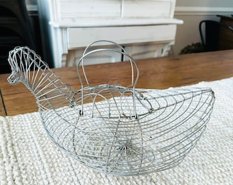 May include: A silver wire chicken-shaped basket with a handle. The basket is sitting on a white woven mat.