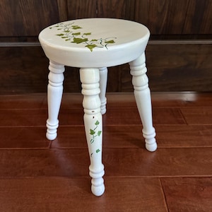 May include: A small, white wooden stool with a round seat and four turned legs. The seat is decorated with green ivy-like designs. The legs also feature green leaf accents. The stool is painted white and stands on a wooden floor.