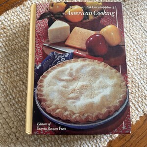 May include: The Illustrated Encyclopedia of American Cooking cookbook with a yellow spine and a red, white, and blue cover featuring a pie, apples, and cheese.
