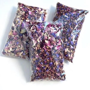 May include: Three clear plastic bags filled with a mixture of dried flower petals in shades of pink, purple, blue, and cream. The petals are small, fragmented, and densely packed within each sealed rectangular pouch, creating a colorful display.