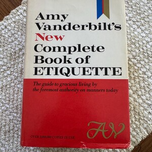 May include: A red hardcover book titled "Amy Vanderbilt's New Complete Book of Etiquette" with gold lettering. The book's subtitle is "The guide to gracious living by the foremost authority on manners today." The bottom of the cover reads "OVER 2,000,000 COPIES IN USE" and has a gold "AV" logo.