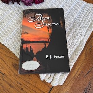 May include: A black paperback book with the title "Bayou Shadows" by B.J. Foster. The cover features a sunset over a bayou with trees and a wooden dock. The book is an autographed copy.