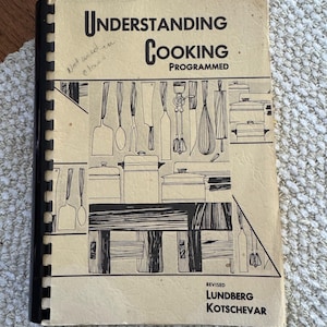 May include: Vintage cookbook titled "Understanding Cooking Programmed" with a black spiral binding. The cover is beige with black text and illustrations of kitchen utensils and cookware. Handwritten note in the upper left corner.