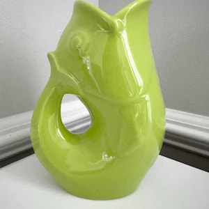 May include: A bright green ceramic pitcher with a fish-like design. The pitcher has a curved handle with an opening and a flared spout. The glossy finish reflects light, highlighting the curves and details of the fish-inspired form. A decorative piece for the home.