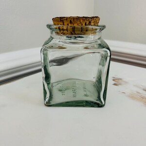 May include: A clear glass jar with a cork lid. The jar is square shaped and has a textured surface. The jar is labeled "MADE IN SPAIN" and "JARRET".