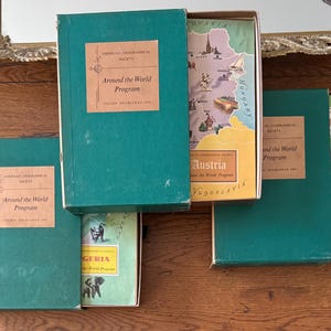 May include: A collection of vintage travel books from the "Around the World Program" series. The books have teal green covers with tan labels. One box is open, revealing a map of Austria and Yugoslavia. The books are arranged on a wooden surface.