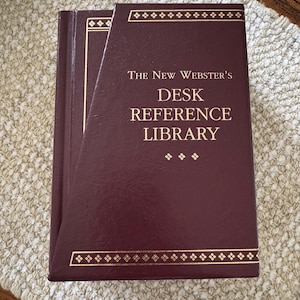 May include: A maroon-colored book titled "The New Webster's Desk Reference Library." The book has gold-colored decorative borders and text. The book is closed and appears to be in good condition.