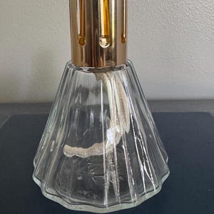 May include: A clear glass fragrance lamp with a gold-colored metal top. The lamp has a flared, conical shape with vertical ridges. A white wick extends from the top. The lamp is designed for aromatherapy and home fragrance.