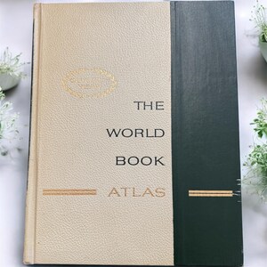 May include: A vintage hardcover book titled "The World Book Atlas." The book has a cream-colored cover with gold lettering and a dark green spine. The cover features the text "The World Book Atlas" and a gold emblem.
