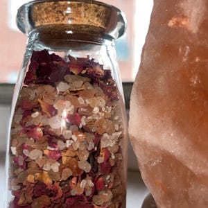 Op de afbeelding: Een heldere glazen pot gevuld met rozenblaadjes en kristallen, afgedekt met een kurken deksel. De pot staat naast een grote, roze Himalaya zoutlamp. De inhoud van de pot is een mix van rode, bruine en heldere elementen.
