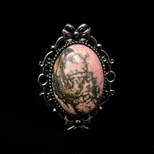 May include: An oval-shaped ring featuring a polished pink and black patterned stone cabochon set within a dark metallic frame. The setting includes intricate scrollwork details and a small bow motif at both the top and bottom of the piece.