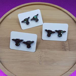 May include: Three pairs of glittery cow-shaped stud earrings on white cards, displayed on a wooden tray against a purple background. The earrings are dark with flecks of green and blue glitter, creating a sparkling effect. The overall theme is jewelry.
