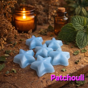Puede incluir: Estrellas de cera azul claro dispuestas sobre una superficie de madera. La palabra "Patchouli" se muestra en texto rosa. Una vela encendida en un frasco de vidrio y una pequeña botella marrón están en el fondo, junto con hojas verdes y hierbas secas.