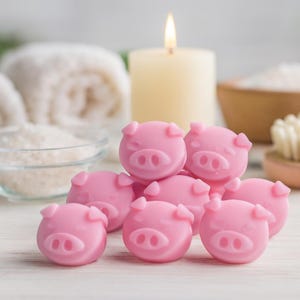 May include: A collection of pink pig-shaped soaps. The soaps are clustered together, with a lit candle and spa-themed items in the background. The soaps have detailed facial features.