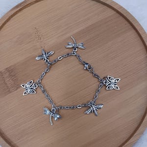 Könnte beinhalten: Silberfarbenes Charm-Armband mit Libellen- und Schmetterlingsanhängern. Das Armband hat eine feine Kette und einen Karabinerverschluss. Die detaillierten Charms verleihen einen Hauch von Natur. Für Handgelenke von ca. 15-20 cm.