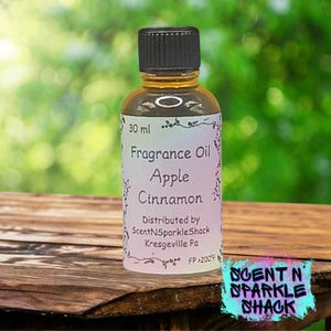 Puede incluir: Una peque&ntilde;a botella de vidrio de aceite de fragancia con tapa negra. La etiqueta dice "Fragrance Oil Apple Cinnamon" y "30 ml". La botella est&aacute; sobre una superficie de madera con un fondo verde borroso. El logotipo "Scent N Sparkle Shack" est&aacute; en la esquina inferior derecha.