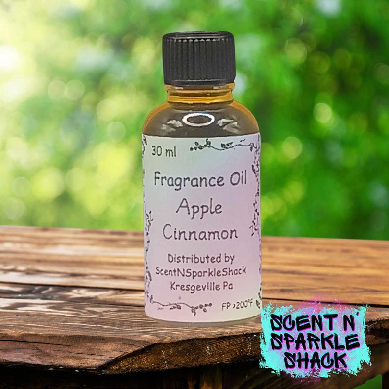 Puede incluir: Una peque&ntilde;a botella de vidrio de aceite de fragancia con tapa negra. La etiqueta dice "Fragrance Oil Apple Cinnamon" y "30 ml". La botella est&aacute; sobre una superficie de madera con un fondo verde borroso. El logotipo "Scent N Sparkle Shack" est&aacute; en la esquina inferior derecha.