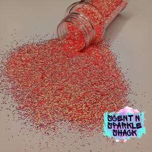 Könnte beinhalten: Ein klarer Plastikbehälter, der umgekippt ist und leuchtend roten Glitzer auf eine weiße Oberfläche verschüttet. Der Glitzer hat eine Mischung aus roten, goldenen und irisierenden Partikeln. Das Bild enthält ein Logo mit der Aufschrift "SCENT N' SPARKLE SHACK".