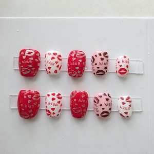 Może przedstawiać: Zestaw dziesięciu sztucznych paznokci o r&oacute;żnych wzorach. Paznokcie mają czerwone, białe i r&oacute;żowe podstawy z wzorami pocałunk&oacute;w i serc. Niekt&oacute;re paznokcie mają wz&oacute;r szminki, a na kilku paznokciach wydrukowano słowo "kiss".