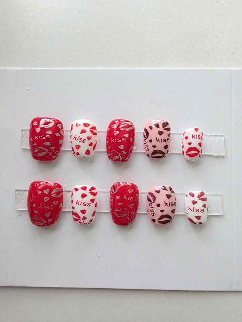 Może przedstawiać: Zestaw dziesięciu sztucznych paznokci o r&oacute;żnych wzorach. Paznokcie mają czerwone, białe i r&oacute;żowe podstawy z wzorami pocałunk&oacute;w i serc. Niekt&oacute;re paznokcie mają wz&oacute;r szminki, a na kilku paznokciach wydrukowano słowo "kiss".