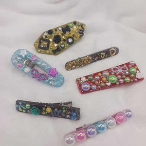 Puede incluir: Una colección de horquillas decorativas en varias formas y colores. Las horquillas presentan adornos como purpurina, perlas y cuentas de colores. Una horquilla tiene un diseño de lazo y estrella, mientras que otra tiene un estampado floral. Sobre un fondo blanco.