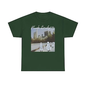 May include: A dark forest green short-sleeve t-shirt featuring a rectangular graphic print on the chest. The print displays a city skyline with several figures dressed in white ghost costumes and the white cursive text Phoebe Bridgers above.