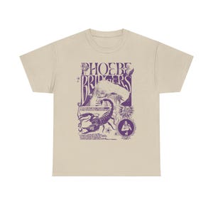 May include: A beige short-sleeve t-shirt featuring a purple graphic print. The design includes the text PHOEBE BRIDGERS, a scorpion illustration, a figure with hands over their eyes, and smaller circular motifs with additional text elements.