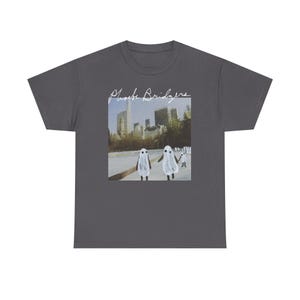 Puede incluir: Una camiseta de manga corta de color gris oscuro con un estampado gráfico central de un horizonte urbano con varias figuras vestidas con disfraces de fantasmas blancos. El texto Phoebe Bridgers aparece en cursiva blanca sobre la imagen.