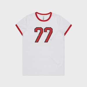Op de afbeelding: Een wit ringer-t-shirt met een rode ronde hals en rode mouwboorden. In het midden van het shirt staat het getal 77 in een vetgedrukt rood lettertype met een zwarte omlijning en een verweerd, versleten textuureffect op de cijfers.