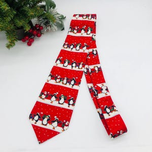 May include: A red necktie with a repeating pattern of dancing penguins on white stripes. The penguins are wearing hats and scarves. The background is red with white polka dots. A festive accessory for the holiday season.