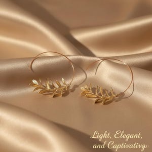 Puede incluir: Pendientes de aro dorados con diseño de hojas. Los pendientes se muestran sobre una superficie de satén. El texto de la imagen dice "Light, Elegant, and Captivating".