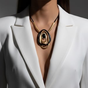 Peut inclure: Un collier doré avec un pendentif noir et doré. Le pendentif a une forme ovale avec des cercles concentriques et une pierre noire au centre. Le collier est porté sur un blazer blanc.