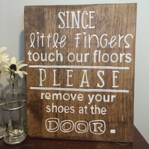 May include: A wooden sign with white text that reads "Since little fingers touch our floors PLEASE remove your shoes at the DOOR."