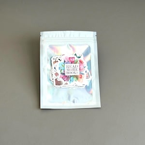 May include: A clear, iridescent zip-lock bag containing a collection of stickers. The stickers feature floral designs and the text "READ MORE BOOKS" in a pink rectangle. The stickers are in various colors, including pink, blue, and purple.