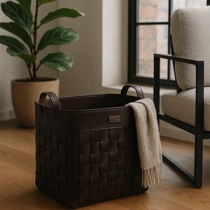 Puede incluir: Una cesta de almacenamiento tejida de color marrón oscuro con dos asas y un ribete de cuero. Una manta beige con flecos está drapeada sobre el lateral. La cesta está sobre un suelo de madera, cerca de una ventana y una planta en maceta.