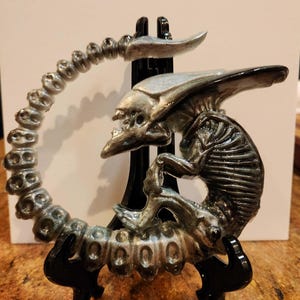 May include: A silver-colored Alien sculpture, featuring a detailed Xenomorph design. The sculpture is mounted on a black stand, showcasing the creature's skeletal structure and elongated head. The tail curves around the body.