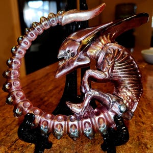 May include: A metallic sculpture of an alien creature in shades of bronze and purple, mounted on a black stand. The sculpture features a detailed, skeletal design with a curved tail and a menacing head. The overall aesthetic is reminiscent of science fiction.