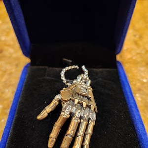 May include: A bronze-toned pendant in the shape of a skeletal hand, with intricate detailing. The pendant is displayed in a blue velvet jewelry box. The hand is made of metal and has a chain attached to it.