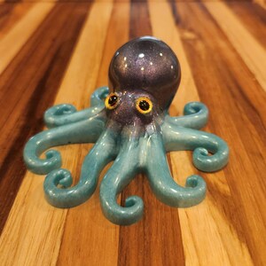 May include: A handcrafted octopus figurine with a dark purple head speckled with white dots and teal tentacles. The octopus has yellow eyes and is set against a wooden background. The figurine is a decorative item.