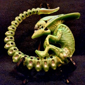 May include: A green alien sculpture with a curved tail and a detailed body. The sculpture has a metallic sheen and is mounted on a black stand. The alien has a detailed head with visible teeth and multiple black holes.