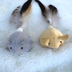 May include: Two mouse-shaped cat toys, one grey and one yellow, with small ears and black eyes. Each toy has feathers in brown, black, and grey attached to the top. The toys are on a white, fluffy surface, resembling a sheepskin rug.