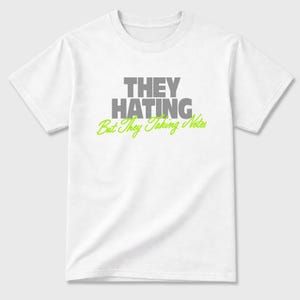 K&ouml;nnte beinhalten: Wei&szlig;es T-Shirt mit dem Text "THEY HATING" in grauen Blockbuchstaben &uuml;ber "But They Taking Notes" in gr&uuml;ner Schrift. Das Shirt hat kurze &Auml;rmel und einen Rundhalsausschnitt.