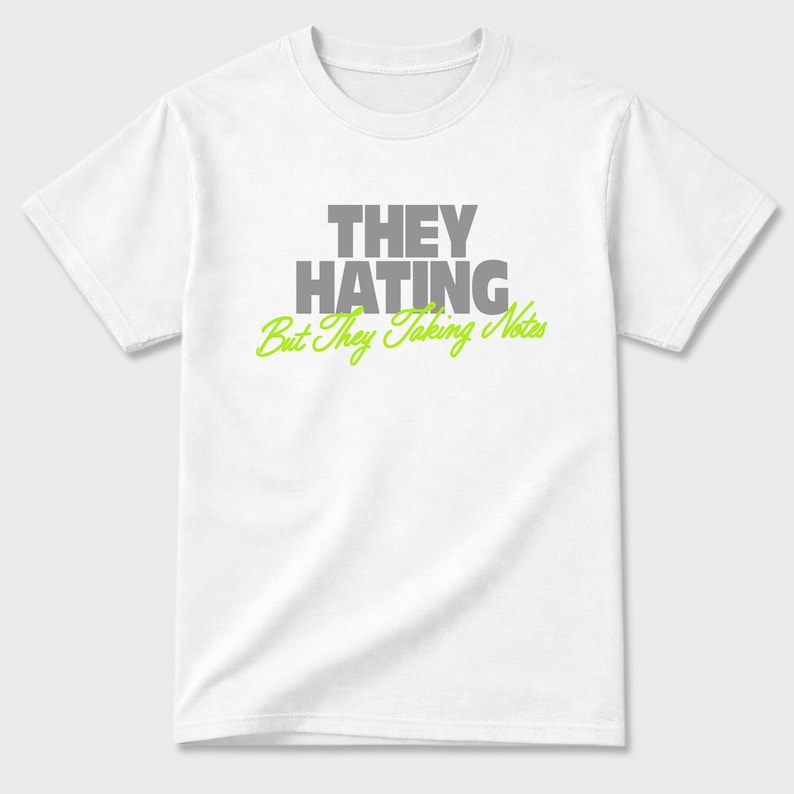 K&ouml;nnte beinhalten: Wei&szlig;es T-Shirt mit dem Text "THEY HATING" in grauen Blockbuchstaben &uuml;ber "But They Taking Notes" in gr&uuml;ner Schrift. Das Shirt hat kurze &Auml;rmel und einen Rundhalsausschnitt.