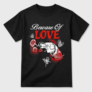 以下が含まれることがあります： 黒いTシャツ。デザインは「Beware Of LOVE」の文字、赤いアクセントの拳銃、バラ、ハートの落書きが特徴です。Tシャツは柔らかい素材で作られています。