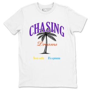 Puede incluir: Camiseta blanca con el texto "CHASING Dreams" en morado y naranja, y un dibujo de palmera negra. Debajo del árbol están las palabras "Never settle" e "It's a process" en amarillo y azul.