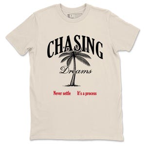 Puede incluir: Una camiseta color crema con las palabras "CHASING Dreams" en negro sobre un gráfico de palmera. Debajo del gráfico, el texto dice "Never settle. It's a process" en rojo.