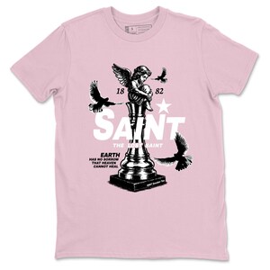 Könnte beinhalten: Hellrosa T-Shirt mit einem schwarz-weißen Grafikdesign. Das Design zeigt einen Engel, Vögel, das Wort "SAINT", die Zahlen "18 82" und den Text "EARTH HAS NO SORROW THAT HEAVEN CANNOT HEAL".