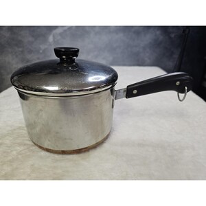 May include: A stainless steel saucepan with a polished finish, featuring a matching lid with a black knob. The pan has a long black handle with a metal hanging loop at the end. The base of the cookware shows a copper-colored bottom surface.