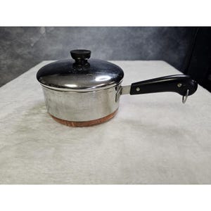 May include: A stainless steel saucepan with a copper-colored base, featuring a matching stainless steel lid with a black knob. The pan has a black handle with a small metal loop at the end for hanging, providing a functional kitchen tool.