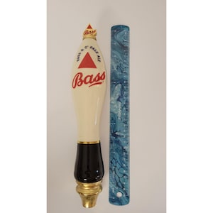 May include: A Bass Pale Ale tap handle with a cream-colored body, black base, and gold accents. The handle features the Bass logo in red script and a red triangle. A blue ruler is next to the tap handle.